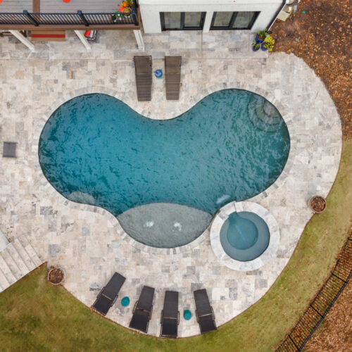 Aerial view of a peanut-shaped swimming pool with a circular hot tub attached on one side. Surrounded by a stone patio, there are lounge chairs and small tables around the pool. The area is bordered by grass and a partial fence.