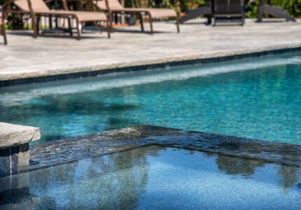 Close-up of a swimming pool with clear, blue water and a stone edge. In the background, there are several lounge chairs on a stone patio, partially shaded by trees.