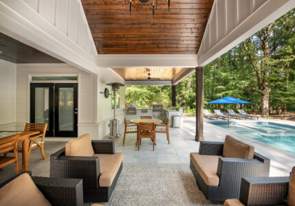 Outdoor patio with wooden ceiling and modern furniture, including a set of wicker chairs and a table. A pool is visible in the background with lounge chairs and a blue umbrella. Surrounded by lush greenery.