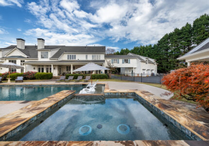 Luxurious backyard featuring a stone-bordered pool with a connected hot tub. Lounge chairs and a striped umbrella sit beside the pool. In the background, theres a large, elegant house with a manicured lawn and tall trees on the right.