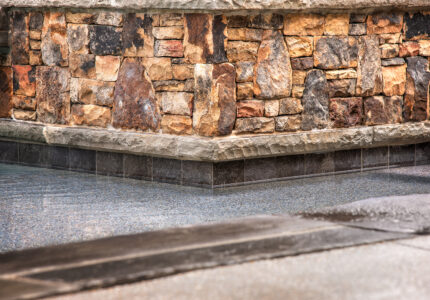 Close-up of a corner of a swimming pool with clear water. The pool is lined with dark tiles and surrounded by a rustic stone wall made of various shades of brown and gray. The edge of the pool is bordered by a stone walkway.