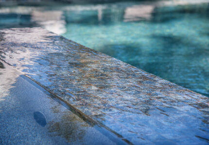 Close-up of a clear blue swimming pool with water gently flowing over an infinity edge. The sunlight reflects off the water, creating shimmering patterns on the surface. A blurred background suggests a peaceful outdoor setting.