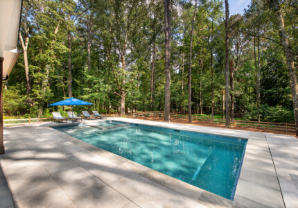 A rectangular swimming pool in a forested backyard, surrounded by tall trees. The pool area has a clean, tiled deck with lounge chairs and a blue umbrella to the left, offering a serene and refreshing atmosphere.