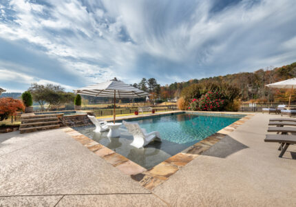 A luxurious outdoor pool with two white lounge chairs partially submerged. The pool is surrounded by stonework and landscaping, including bushes and trees. There are several shaded sitting areas, and a scenic view of hills under a partly cloudy sky.