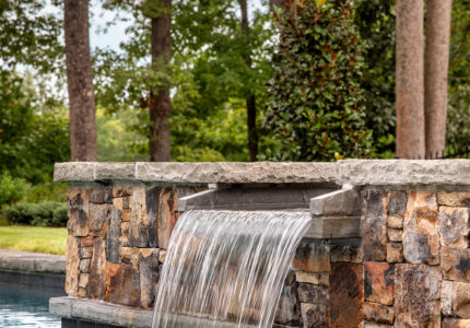 Stone waterfall feature with water cascading into a pool, surrounded by lush green trees and foliage in the background. The scene is serene and picturesque, creating a tranquil outdoor setting.