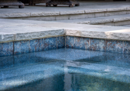 Close-up of a corner of a swimming pool showing clear, blue water and stone-tiled edges. The surface of the pool reflects the tiles, and steps lead upward in the background, creating a serene and inviting outdoor setting.