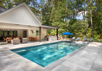 A modern backyard features a rectangular swimming pool surrounded by a stone patio. There are several lounge chairs with a blue umbrella beside the pool. A covered outdoor dining area and grill are adjacent to a lush green forest.