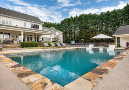 Luxurious backyard with a large, rectangular swimming pool surrounded by a stone deck. Several lounge chairs and an umbrella are placed around the pool. A modern house with a patio and a row of trees are in the background under a partly cloudy sky.