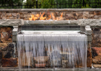 A modern outdoor water feature with water cascading over stone ledges into a pool. In the background, a rectangular fire pit is lit, surrounded by a stone wall. Lush greenery is visible beyond the wall.