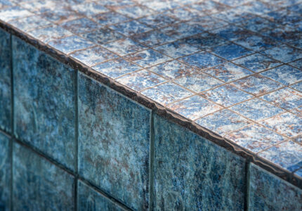 Close-up of a tiled surface featuring small square tiles in various shades of blue with a textured, weathered appearance. The tiles are arranged neatly, with a visible edge marking the boundary between horizontal and vertical surfaces.