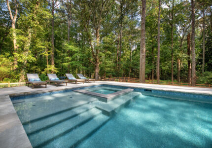 A serene outdoor pool surrounded by lush trees. Four lounge chairs are aligned on the left-hand side of the pool. Sunlight filters through the tall trees, casting soft shadows on the turquoise water. A wooden fence runs along the background.