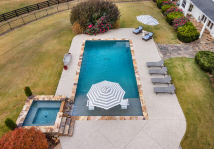 Aerial view of a backyard featuring a rectangular pool with striped umbrellas, surrounded by a stone patio. Several lounge chairs are placed nearby. A smaller jacuzzi is adjacent to the pool, set in a spacious green lawn with shrubs along the sides.