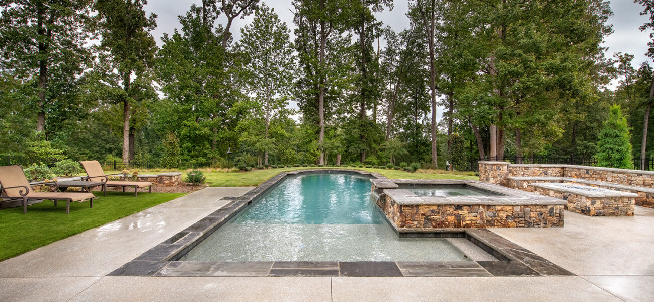 A rectangular pool with clear water is surrounded by a stone patio. There are two loungers on the grass to the left and a spa area to the right. Tall trees form a lush backdrop under a cloudy sky.