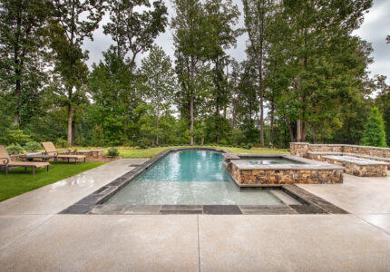 A rectangular pool with clear water is surrounded by a stone patio. There are two loungers on the grass to the left and a spa area to the right. Tall trees form a lush backdrop under a cloudy sky.