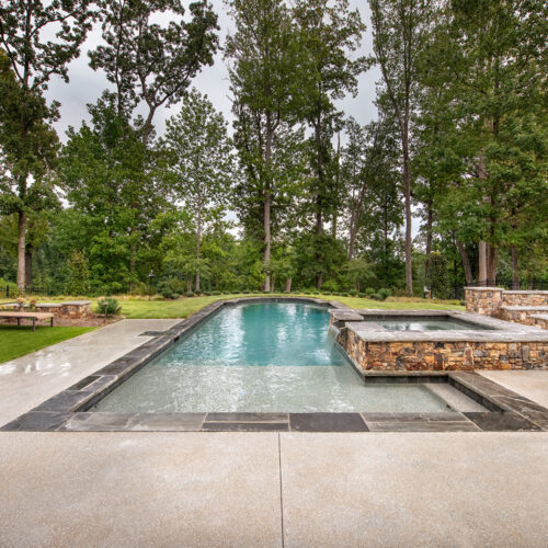 A rectangular pool with clear water is surrounded by a stone patio. There are two loungers on the grass to the left and a spa area to the right. Tall trees form a lush backdrop under a cloudy sky.