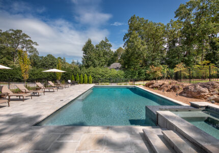 A luxurious outdoor pool area with a modern rectangular pool and attached hot tub. Sun loungers and umbrellas line the stone-tiled patio. Lush green trees and shrubs surround the space, with a clear blue sky above.