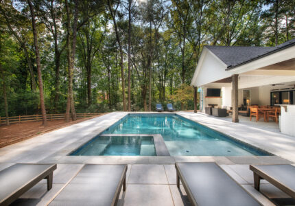 Outdoor pool area with a rectangular pool surrounded by lounge chairs. A small waterfall feature is in the foreground. Beyond the pool, a covered patio with furniture is visible, and trees create a wooded backdrop.