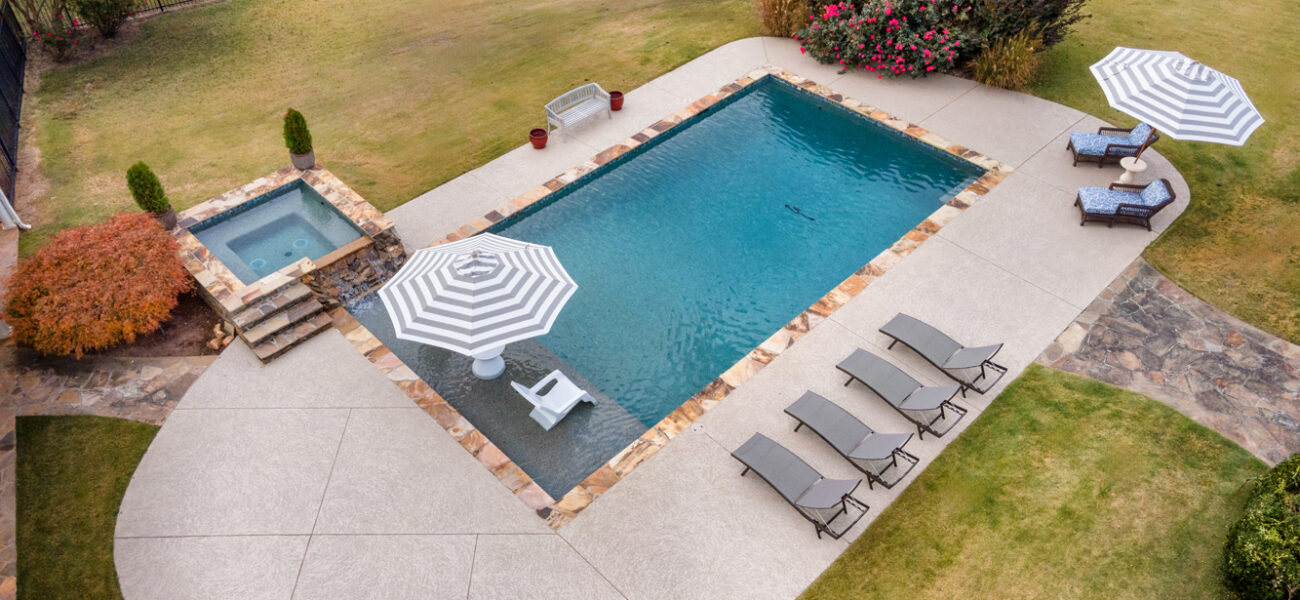 Aerial view of a backyard with a rectangular pool and adjacent hot tub. Two striped umbrellas provide shade over lounge chairs. Paved pathways surround the pool, with a grassy lawn and wooden fence in the background. Shrubs and a tree complete the scene.