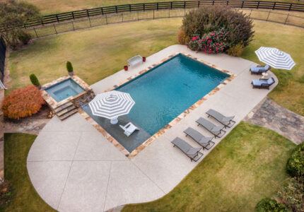 Aerial view of a backyard with a rectangular pool and adjacent hot tub. Two striped umbrellas provide shade over lounge chairs. Paved pathways surround the pool, with a grassy lawn and wooden fence in the background. Shrubs and a tree complete the scene.