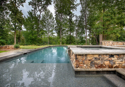 A serene outdoor swimming pool surrounded by lush trees and greenery. The pool features a stone wall with a waterfall, and there is a paved area with natural stone accents. The sky is overcast, creating a tranquil atmosphere.