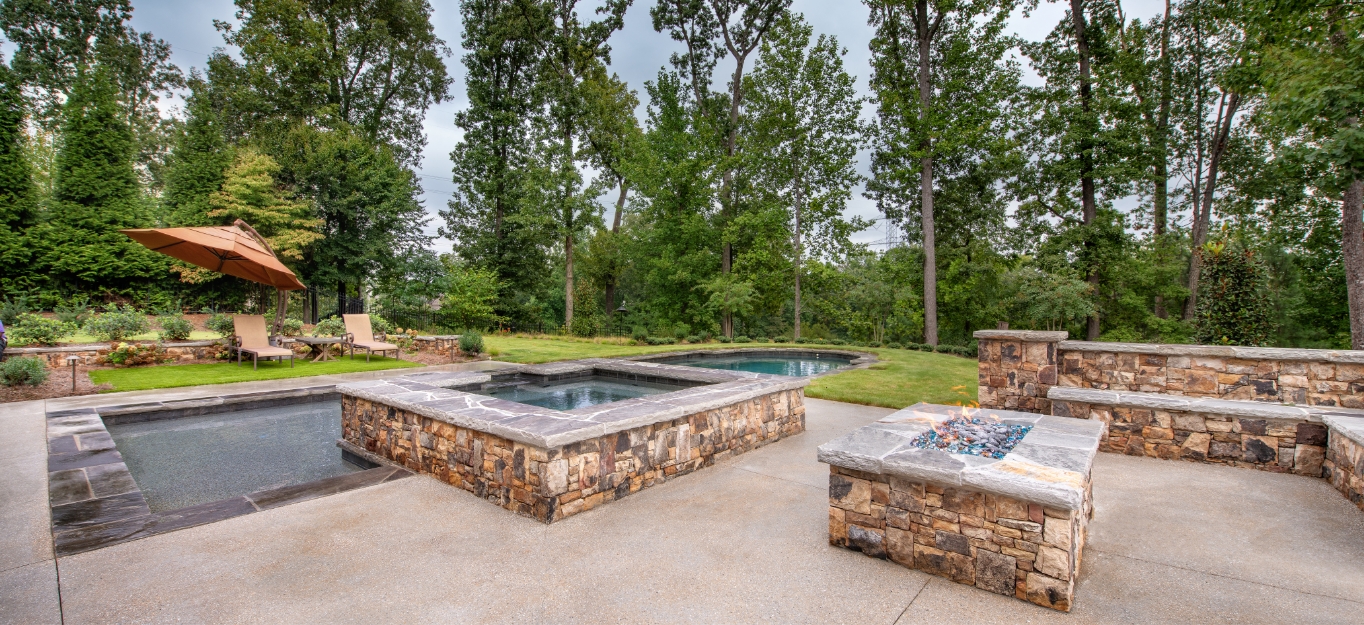 A spacious outdoor patio features a hot tub and pool surrounded by stone accents. Theres a stone fire pit in the foreground and lounge chairs with an umbrella in the background, set against a backdrop of tall trees.