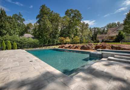 A rectangular outdoor pool surrounded by a stone deck, set in a landscaped yard with trees and bushes. Theres a small waterfall feature with rocks on one side, and steps leading into the pool on the other. A fence and neighboring house are visible.