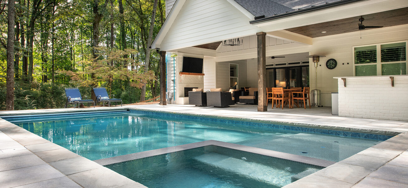 Outdoor swimming pool with integrated hot tub in the foreground, surrounded by a stone deck. In the background, theres a covered patio with outdoor seating, dining set, and a TV. Lush trees surround the area, providing a serene backdrop.