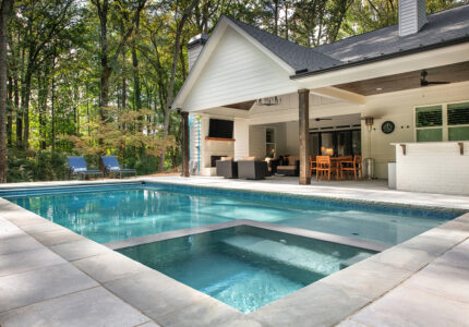 Outdoor swimming pool with integrated hot tub in the foreground, surrounded by a stone deck. In the background, theres a covered patio with outdoor seating, dining set, and a TV. Lush trees surround the area, providing a serene backdrop.