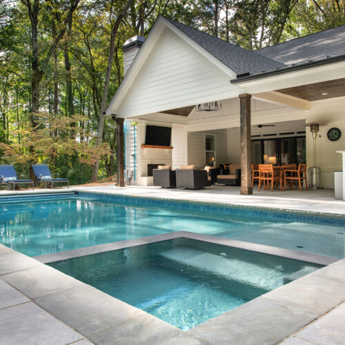 Outdoor swimming pool with integrated hot tub in the foreground, surrounded by a stone deck. In the background, theres a covered patio with outdoor seating, dining set, and a TV. Lush trees surround the area, providing a serene backdrop.