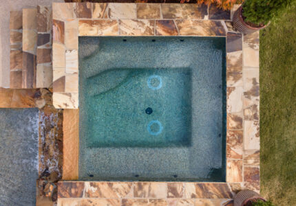 Aerial view of a square hot tub with textured stone tiles around the edges. The water is clear, showing two circular jets at the bottom. Steps leading down into the tub are visible on the left. The surrounding area includes grass and planters.