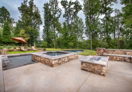 An outdoor space features a stone hot tub, a pool, and a stone seating area with a fire pit. Trees and greenery surround the area, contributing to a serene atmosphere. Two lounge chairs with an umbrella sit in the background.