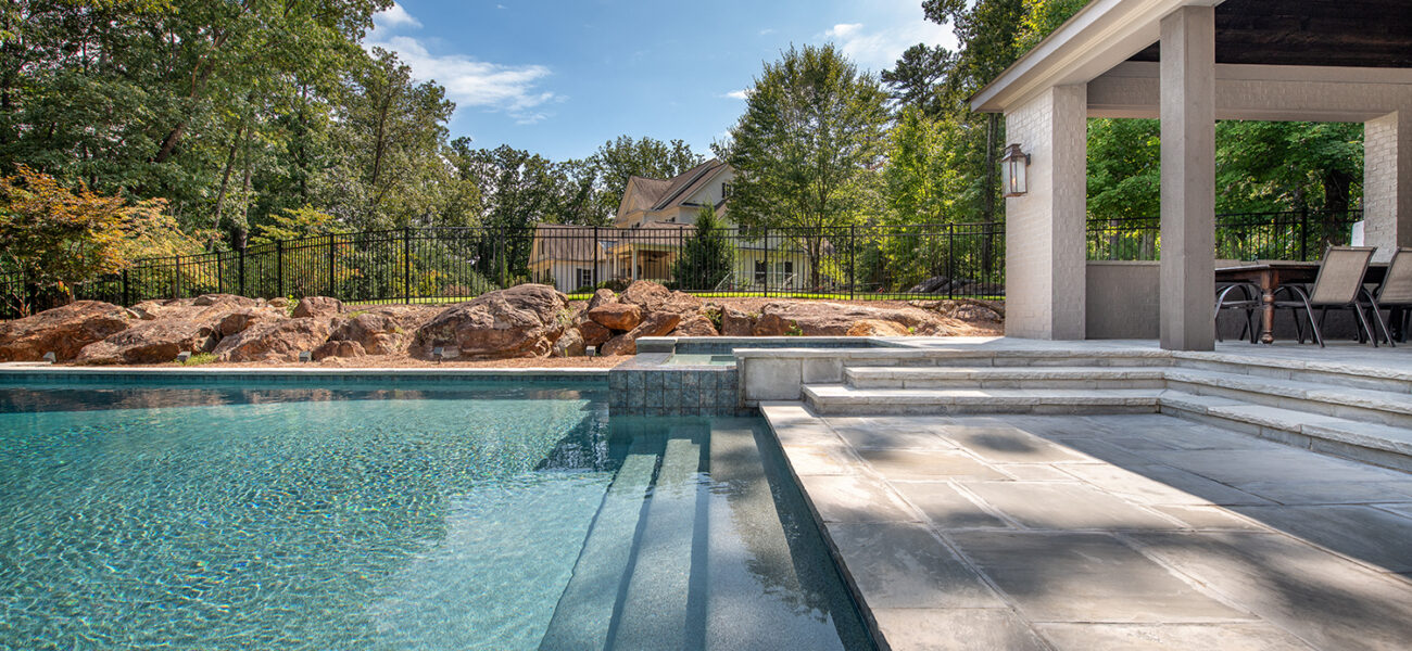 View of a luxurious backyard pool with large stone steps leading into the water. A modern, covered patio area with chairs is on the right. Trees and rocks surround the area, with a house and fence visible in the background under a clear sky.