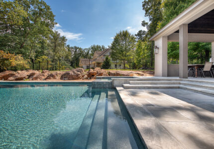 View of a luxurious backyard pool with large stone steps leading into the water. A modern, covered patio area with chairs is on the right. Trees and rocks surround the area, with a house and fence visible in the background under a clear sky.