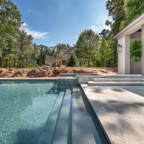 View of a luxurious backyard pool with large stone steps leading into the water. A modern, covered patio area with chairs is on the right. Trees and rocks surround the area, with a house and fence visible in the background under a clear sky.