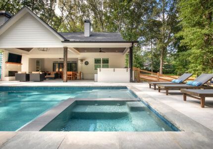 A spacious backyard features a modern house with a covered patio. The patio has outdoor furniture and a wall-mounted TV. A large swimming pool with a connected hot tub is in the foreground, surrounded by a stone deck and trees in the background.