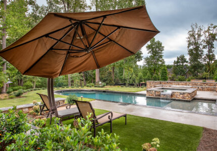 A luxurious backyard with a large swimming pool and a hot tub, surrounded by trees and a garden. Two lounge chairs sit under a large, brown patio umbrella on a grassy area, offering a relaxing view of the serene setting.