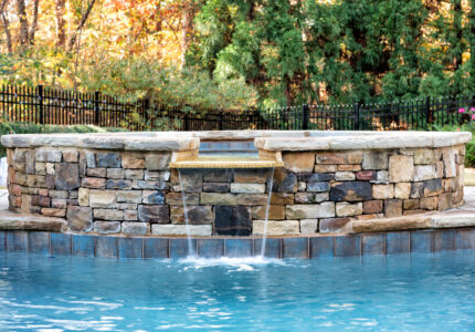 A stone-bordered hot tub overflows into a swimming pool below. The water cascades down the stone wall into the clear blue pool. In the background, there is a black fence, greenery, and autumn trees with colorful leaves.