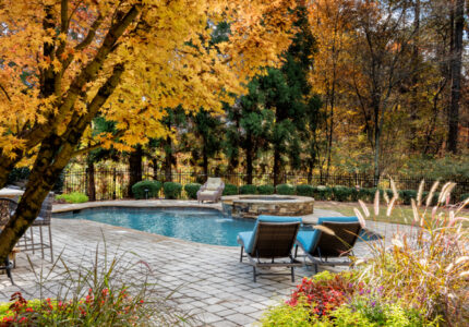 Poolside area with two lounge chairs set on a patio, surrounded by vibrant autumn trees. A stone path with colorful flowers and shrubs leads toward the pool, creating a serene and inviting atmosphere.