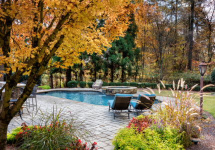 A serene backyard features a swimming pool surrounded by patio chairs, lush greenery, and colorful autumn foliage. Tall trees provide a backdrop, with a lamp post adding charm to the tranquil setting.