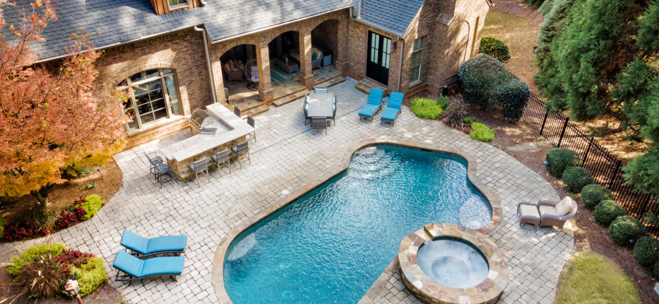 Aerial view of a backyard with a pool and spa. The stone patio area includes lounge chairs and a shaded seating area near the house, which is a brick building with a gray roof. Trees and shrubs border the yard.