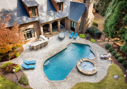 Aerial view of a backyard with a pool and spa. The stone patio area includes lounge chairs and a shaded seating area near the house, which is a brick building with a gray roof. Trees and shrubs border the yard.