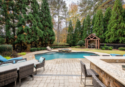 A backyard features a kidney-shaped pool with a hot tub, surrounded by patio furniture and lounge chairs. Tall, green pine trees border the area, and a wooden gazebo is in the background, all under a partly cloudy sky.