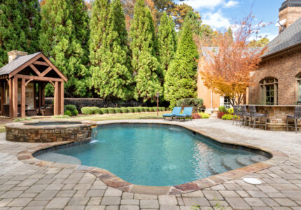 Outdoor pool area surrounded by stone tiles, a brick building, and tall trees. The pool has a curved shape, with lounge chairs nearby and a wooden gazebo in the background under a partly cloudy sky.