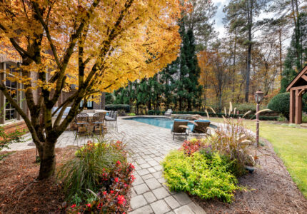 A backyard scene with a cobblestone patio featuring outdoor furniture and a pool. A vibrant yellow tree and lush greenery surround the area. Theres a wooden pergola on the right and tall evergreens in the background.