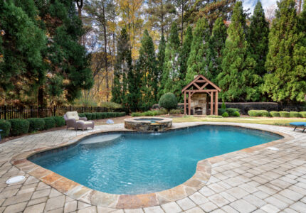 A backyard features a large, kidney-shaped pool surrounded by a stone-tiled patio. There are lounge chairs and an outdoor seating area. Tall evergreen trees and a small wooden gazebo with a fireplace are in the background.