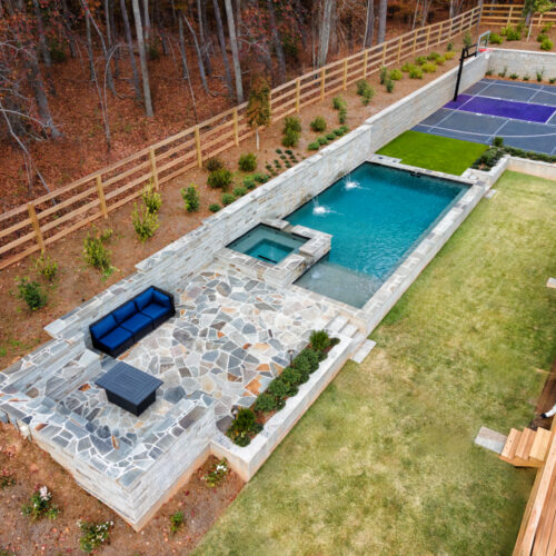 Aerial view of a backyard featuring a stone patio with a blue couch and table, a rectangular pool and spa, a small grassy area, and a basketball court. The area is fenced, bordered by trees and shrubs.